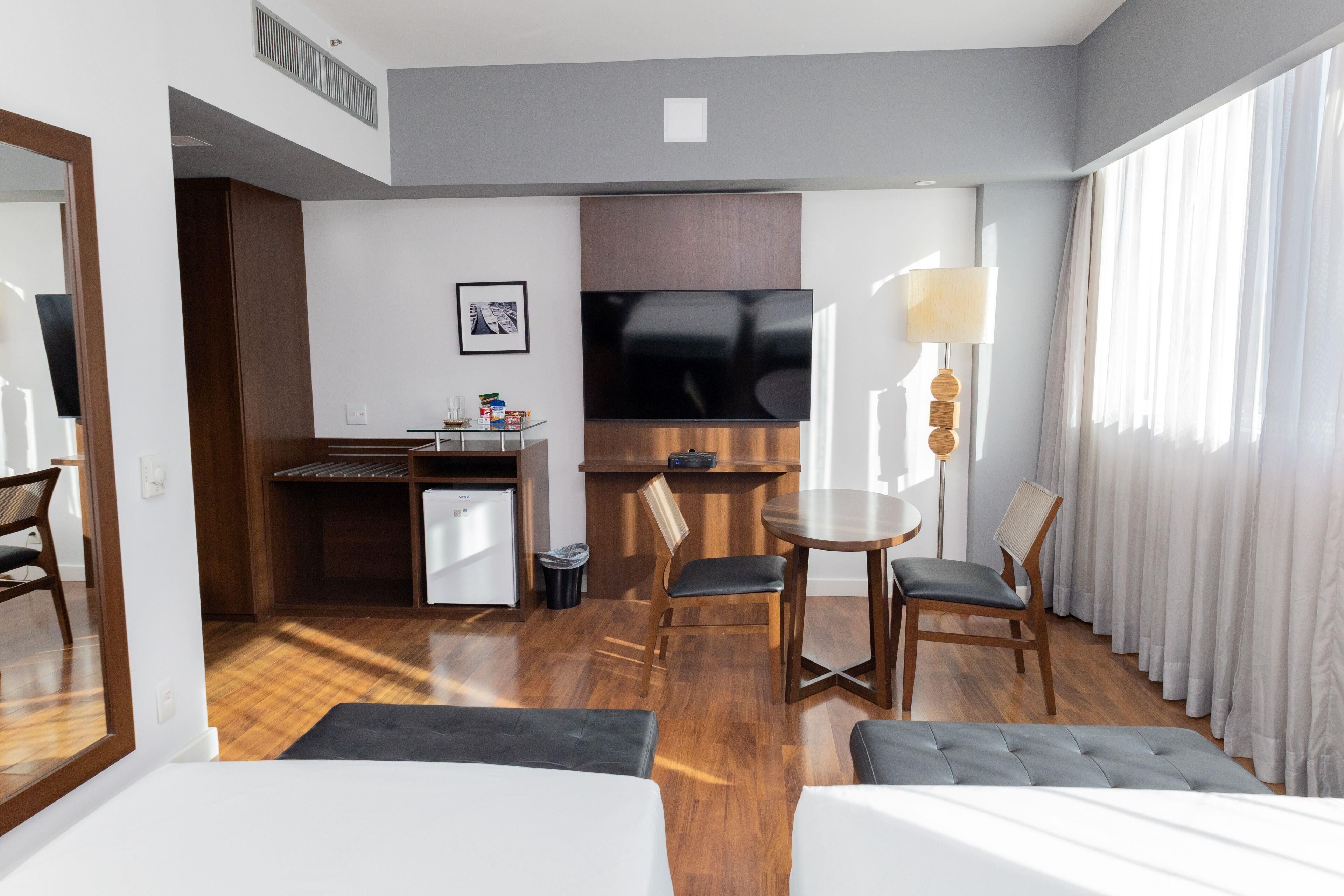 Una habitación de hotel con cama, escritorio, silla, televisión y ventana con cortinas.