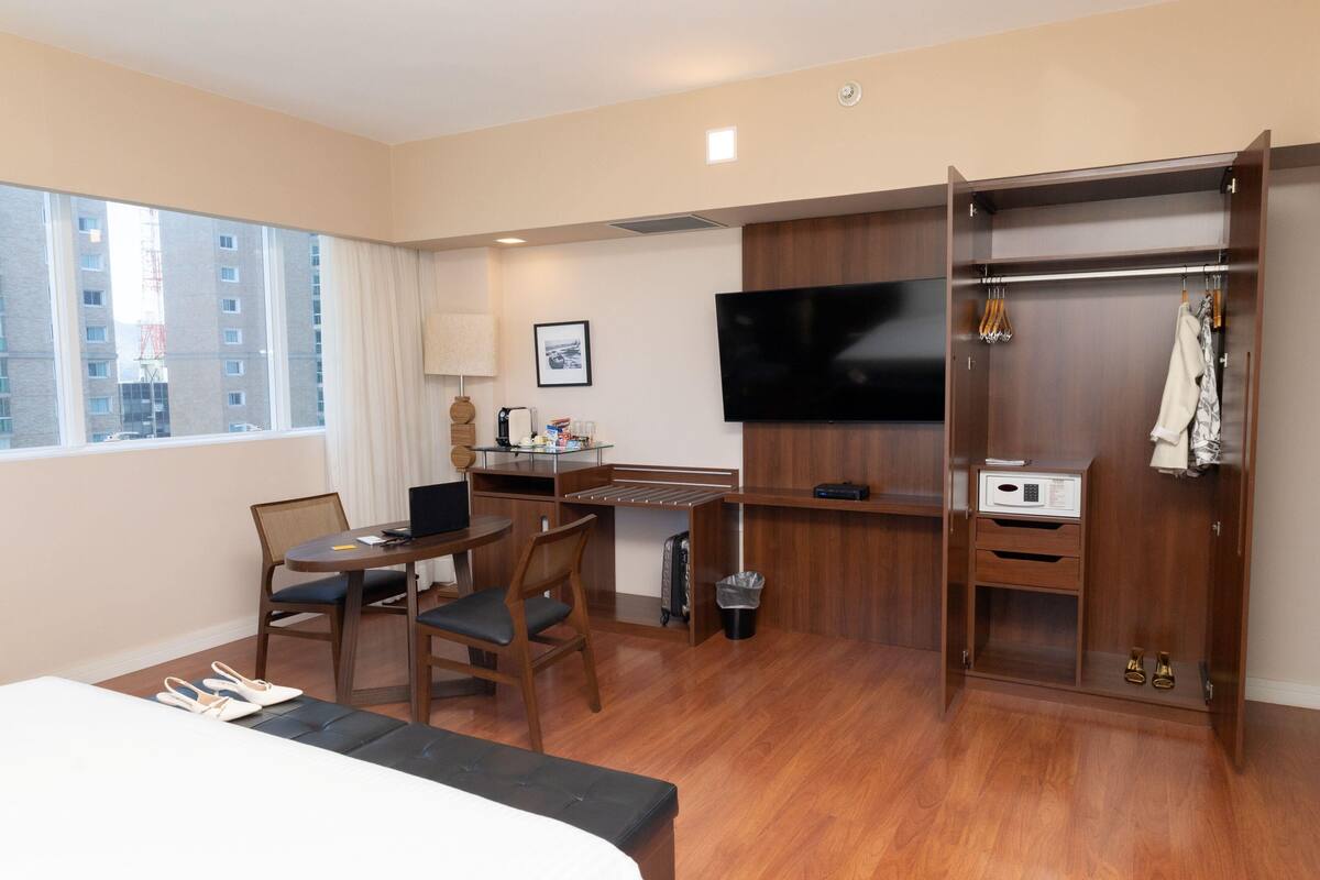 A hotel room with a bed, a desk with a computer, a TV, a wardrobe, and a window with a view of buildings.
