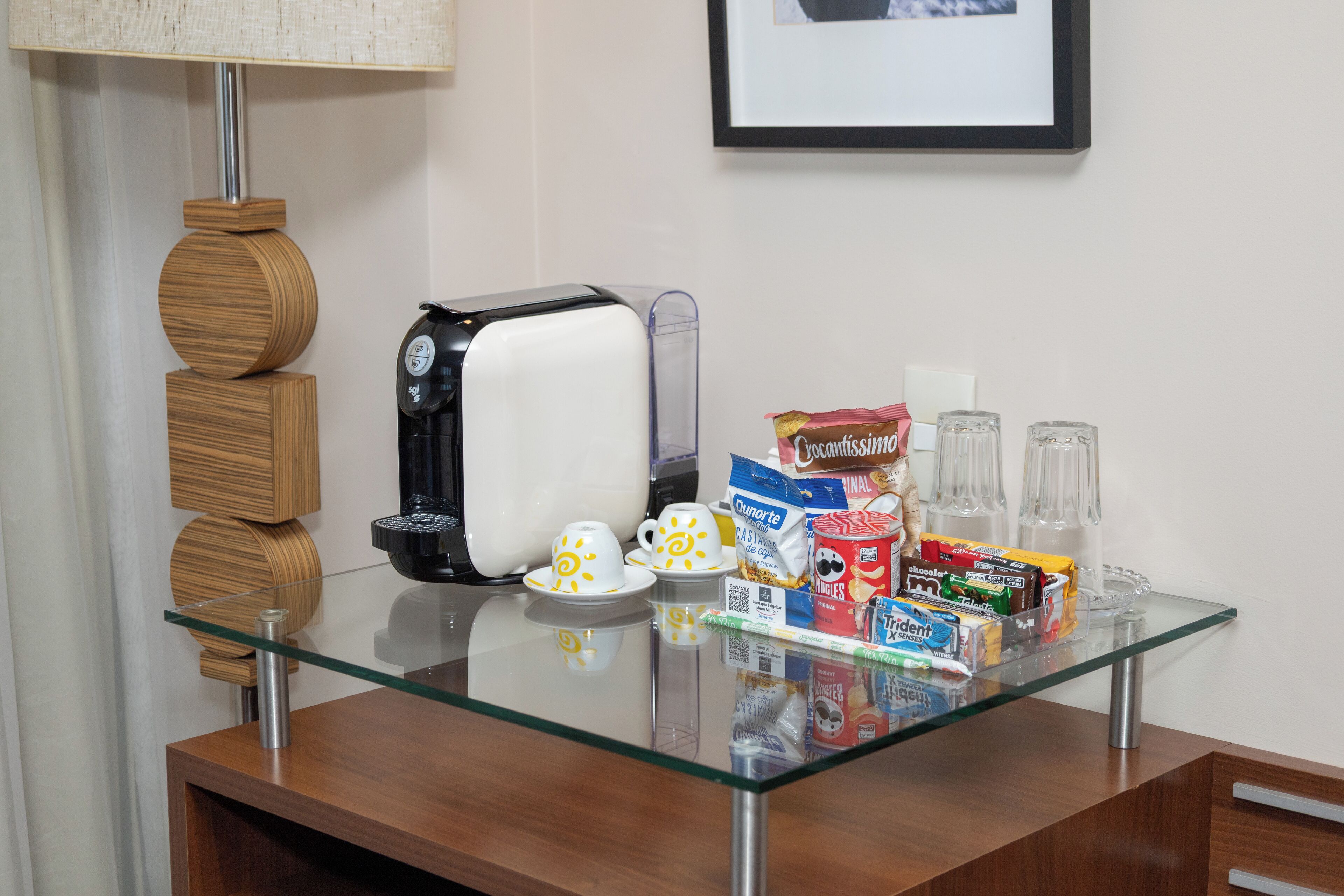 Una mesa con cubierta de vidrio, una tostadora, tazas blancas, una cafetera y diversos alimentos.