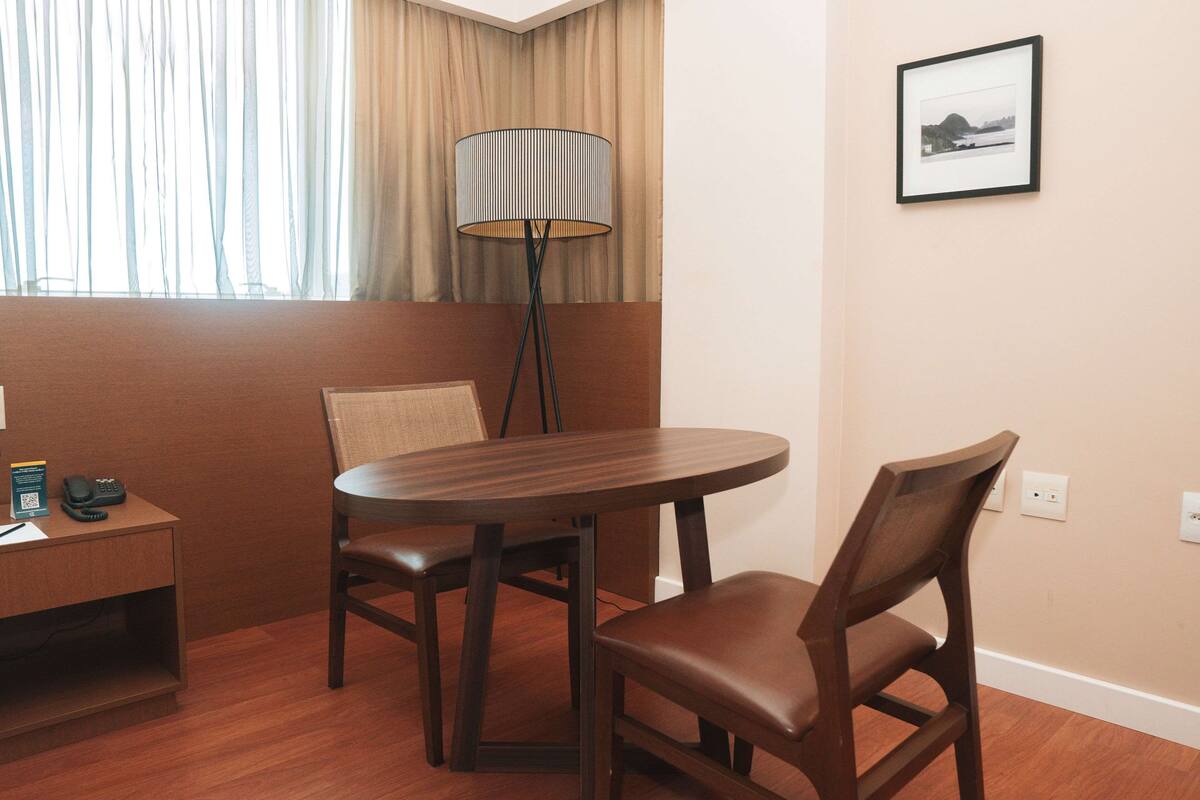 A hotel room with a round wooden table, two chairs, a floor lamp, a small desk with a telephone and a brochure, and a framed picture on the wall.