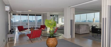 A modern living room with a glass coffee table, red chairs, and a large window offering a city view.