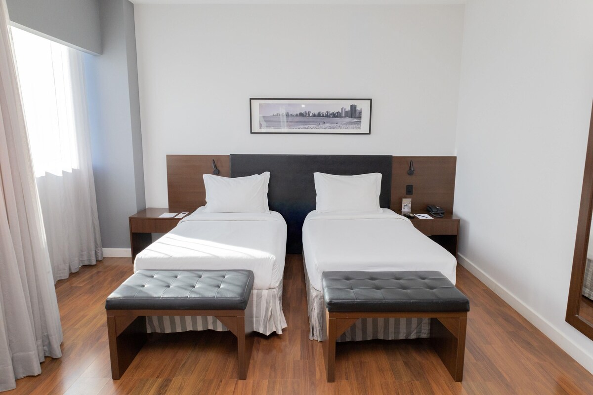 A hotel room with two beds, a cityscape picture on the wall, and a window with curtains.