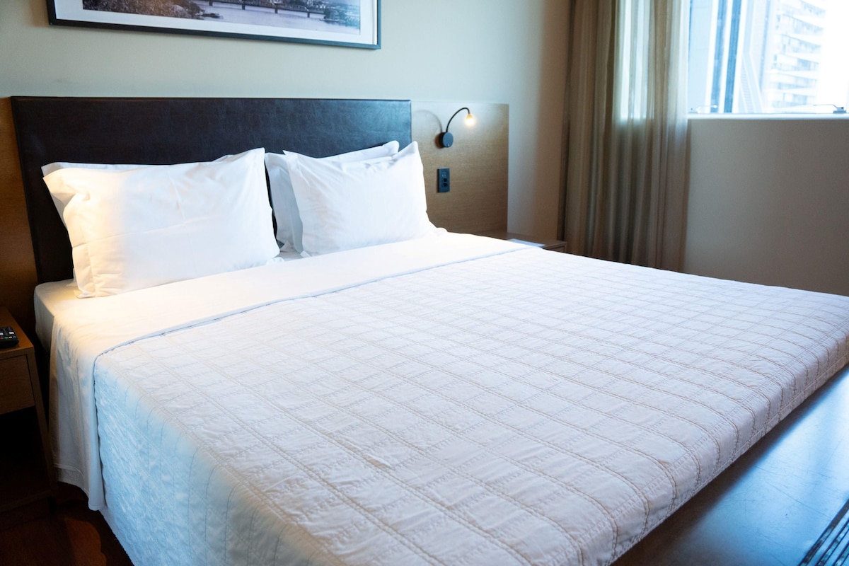 A neatly made bed with white linens, a dark headboard, and a wall-mounted light fixture.