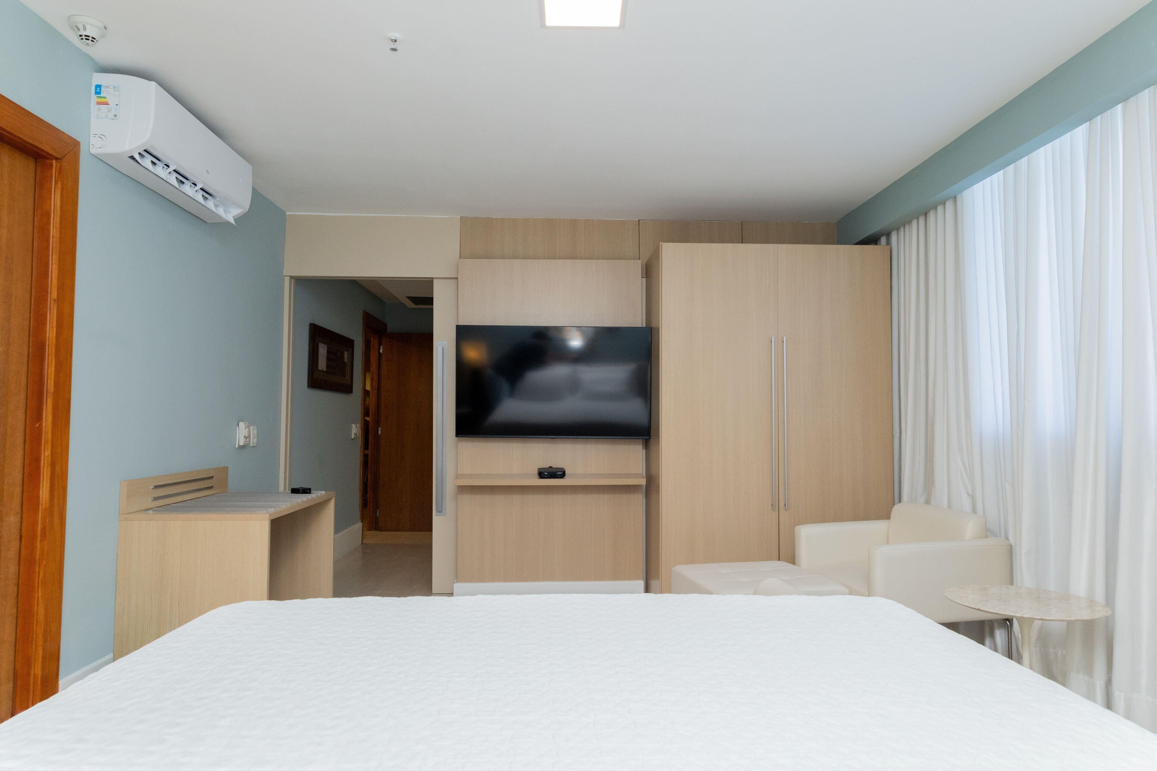 A hotel room with a bed, a TV mounted on the wall, a desk, a chair, and a small table.