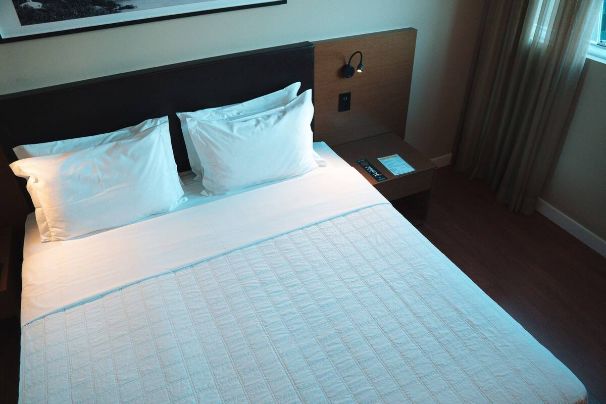 A neatly made bed with white linens and pillows, a bedside table with a lamp, and a framed picture on the wall.