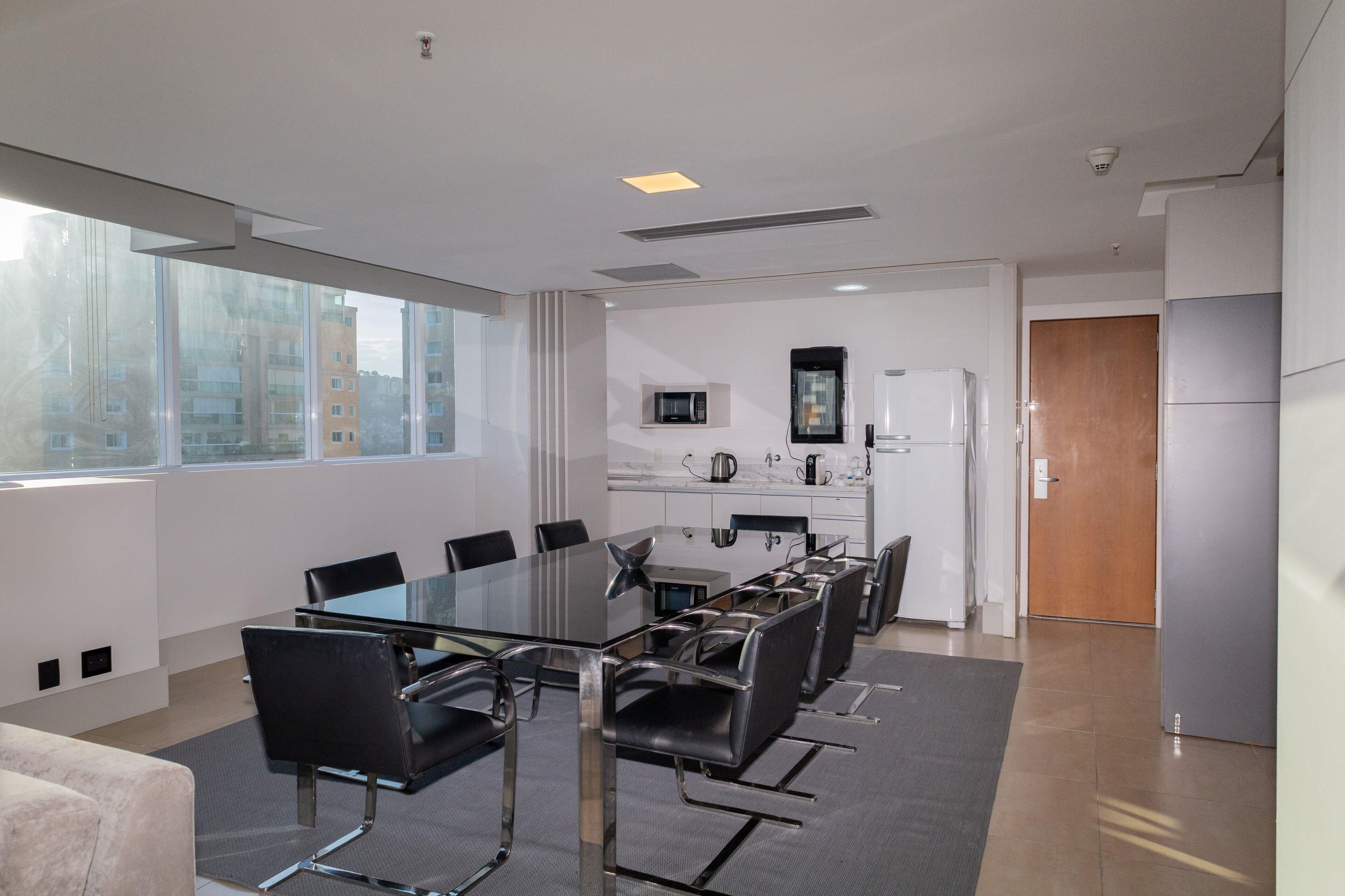 A modern conference room with a glass table and black chairs, a kitchenette with a microwave and refrigerator, and large windows offering a city view.