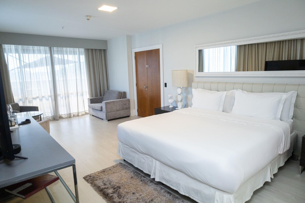 A hotel room with a large bed, a desk, a chair, a TV, and a window with curtains.