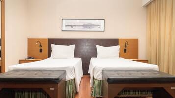 Quarto de hotel com duas camas, cabeceira de madeira, abajures, quadro emoldurado na parede e janela com cortinas.