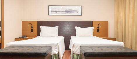 A hotel room with two beds, a wooden headboard, bedside lamps, a framed picture on the wall, and a window with curtains.