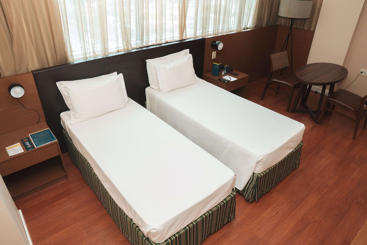 A hotel room with two single beds, a desk, a chair, a lamp, and a small table.
