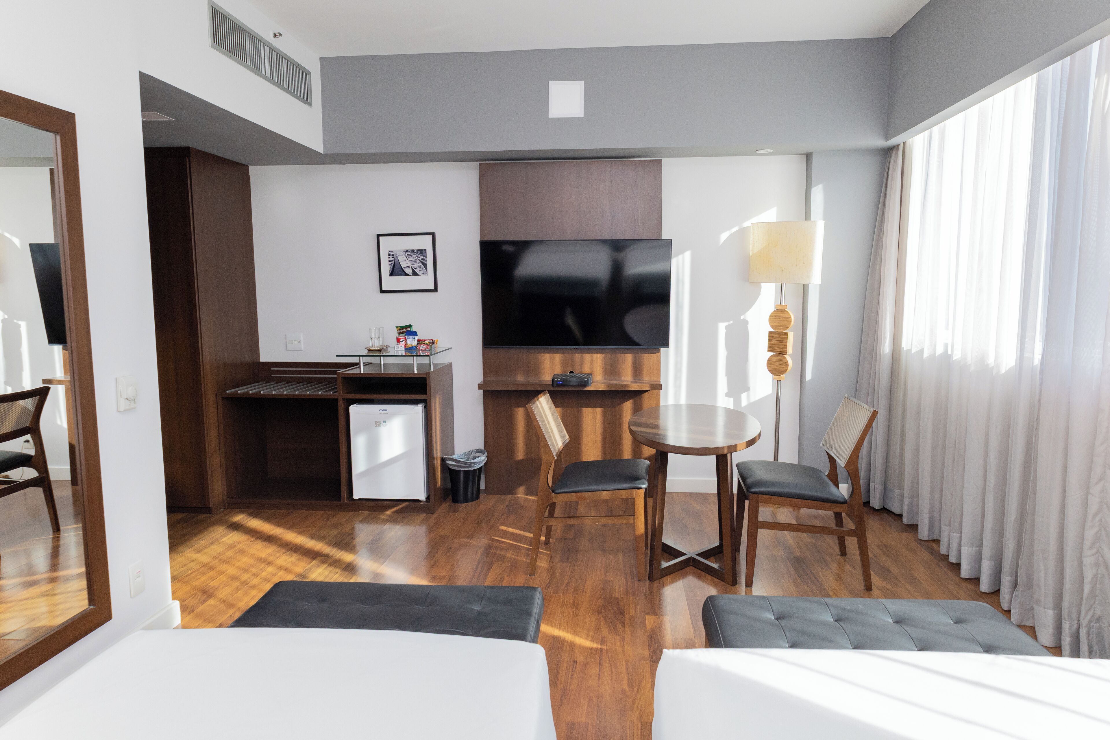 A hotel room with a bed, a desk with a chair, a small kitchenette, a TV, and a window with curtains.