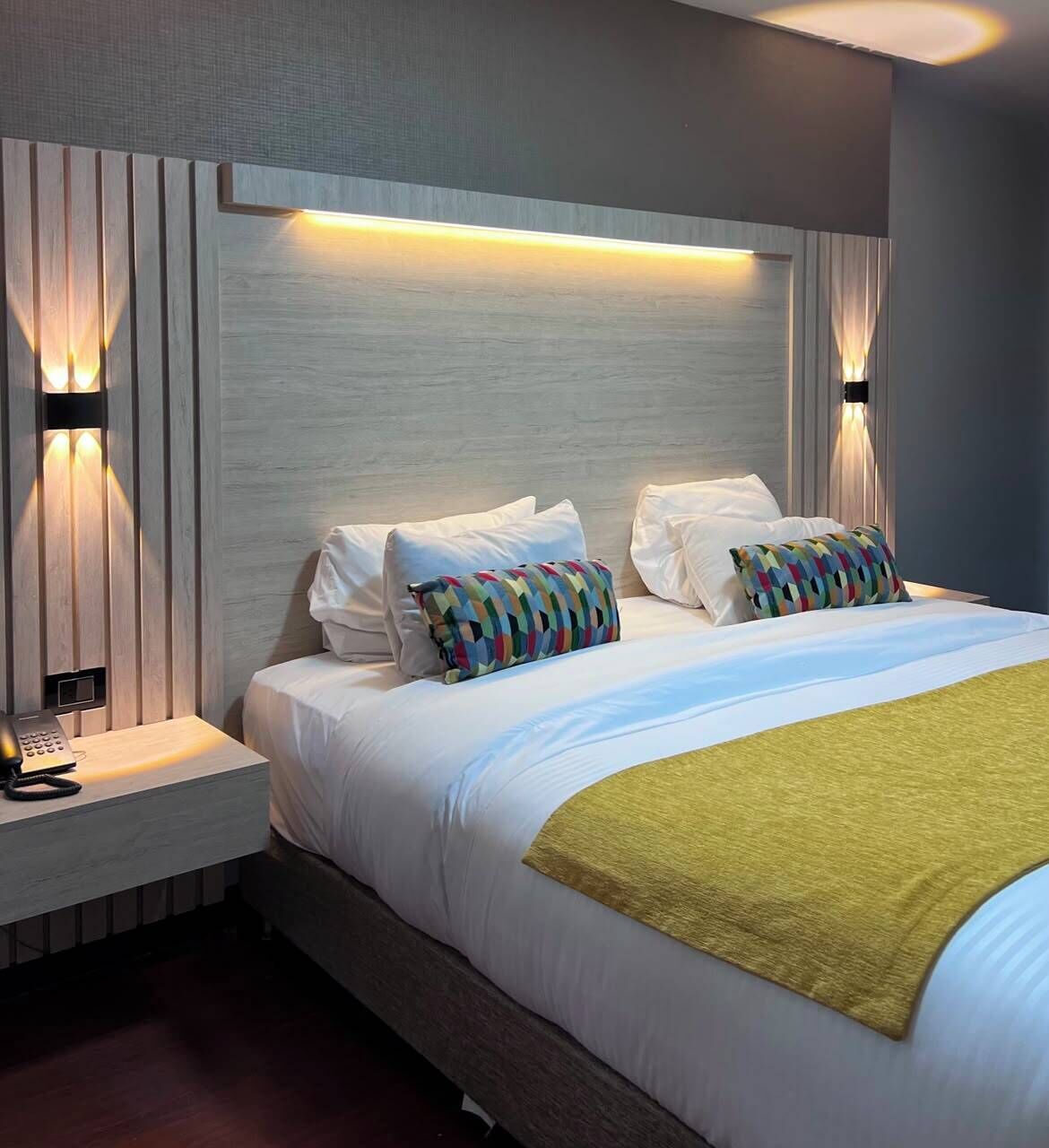 Una habitación de hotel moderna con cama, ropa de cama blanca y una manta verde, además de dos lámparas de noche.