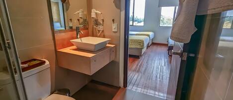 A modern bathroom with a white sink, a mirror, and a toilet. There is a bed visible in the adjacent room.