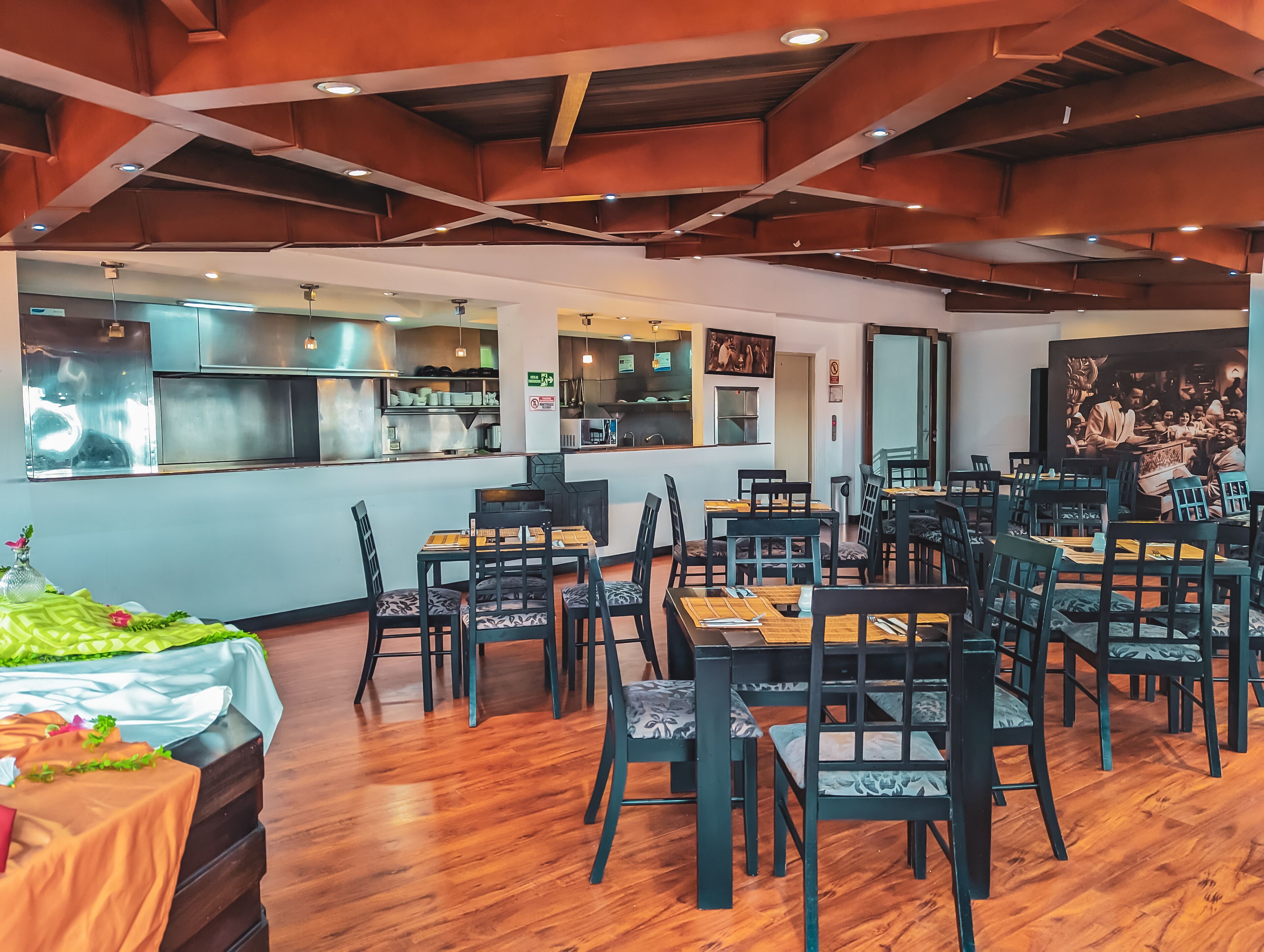 Un restaurante con suelo de madera, sillas negras y mesas. Se observa una zona de cocina con electrodomésticos de acero inoxidable y una barra.