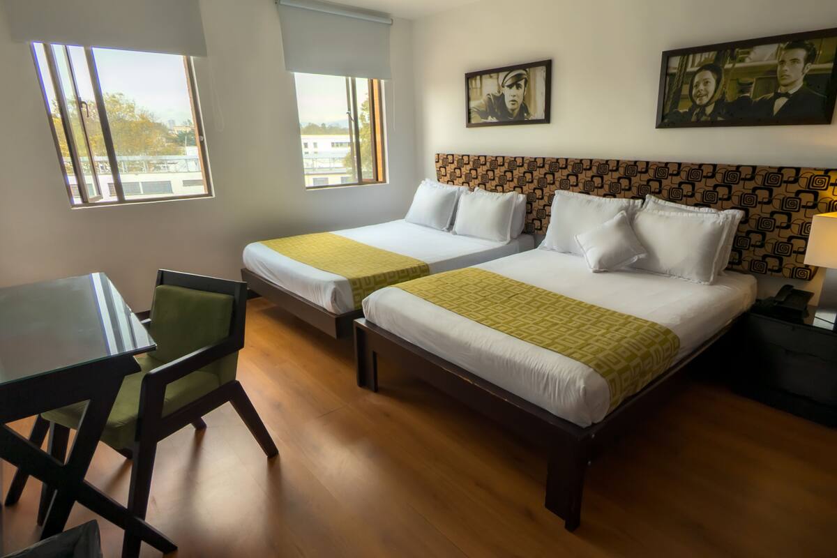 Habitación de hotel con dos camas individuales, un escritorio, una silla, una lámpara y dos cuadros en la pared.