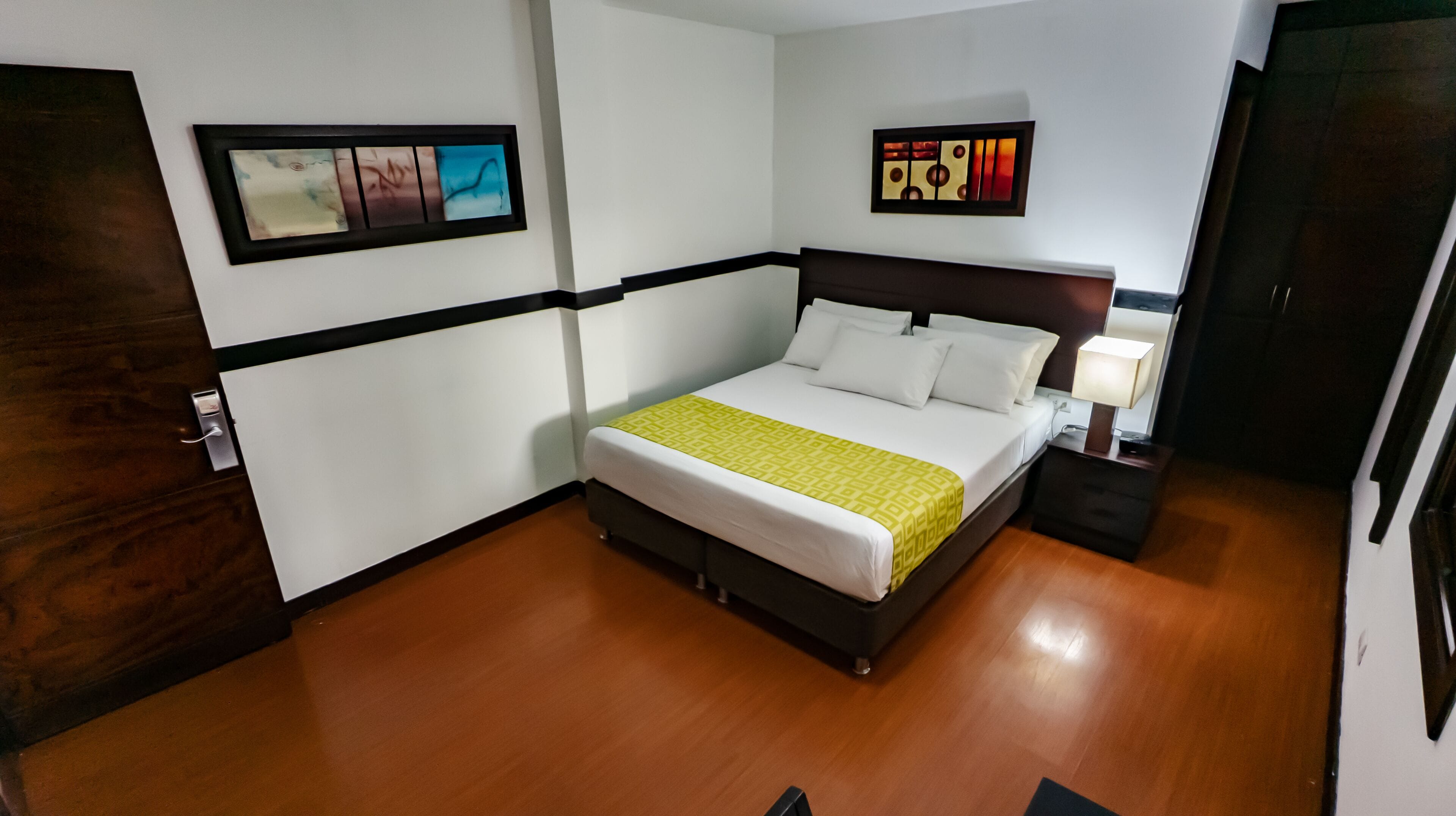 A hotel room with a bed, bedside table, lamp, artwork on the walls, and wooden flooring.
