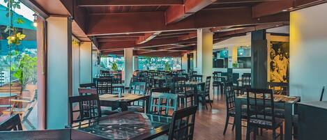 A spacious restaurant with high ceilings, wooden beams, and large windows offering views of the city. Tables and chairs are arranged for dining.