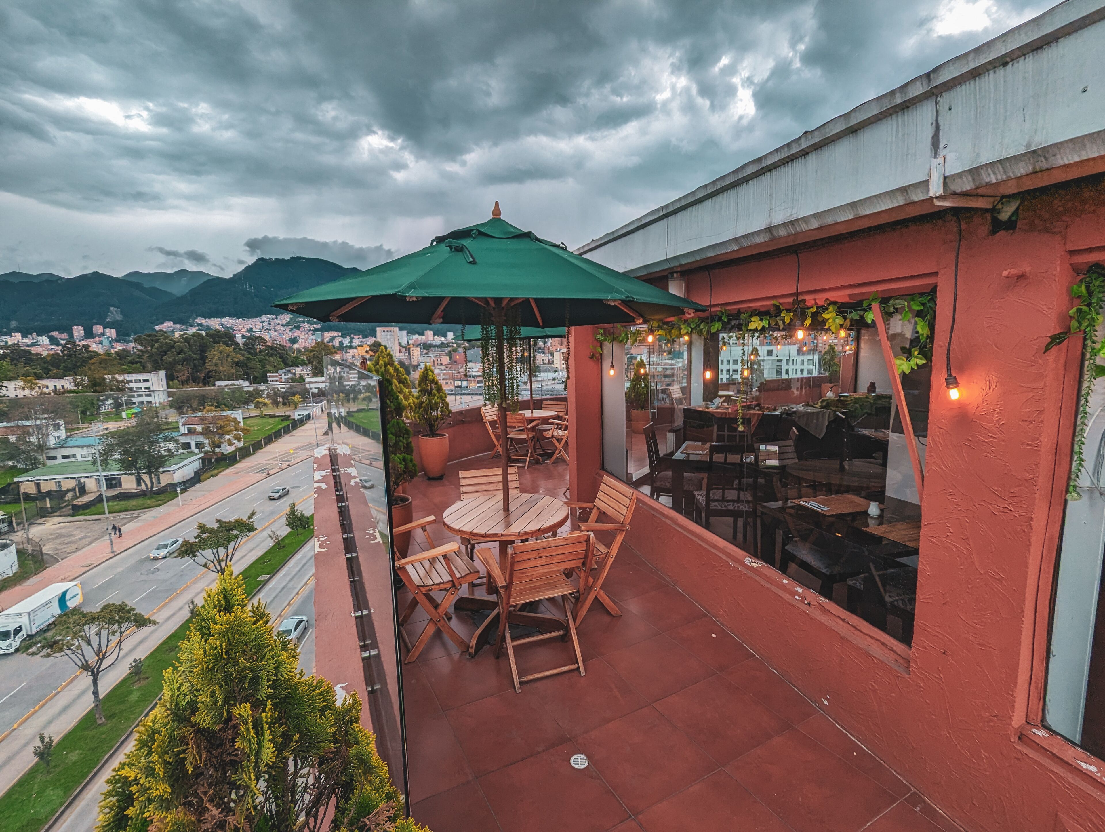 A balcony with a table and chairs, a green umbrella, and a view of a cityscape with mountains in the background.