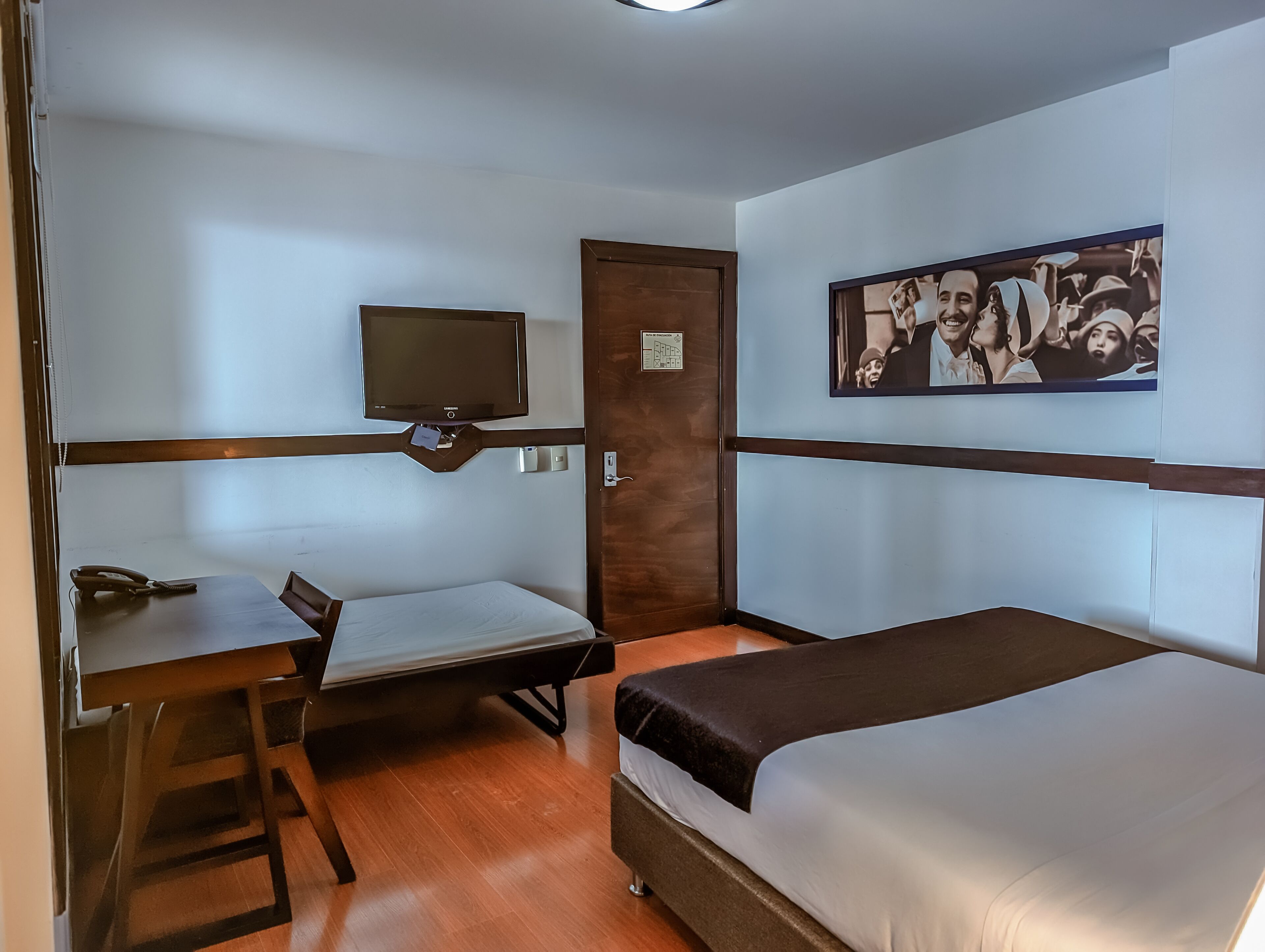 Ein kleines Hotelzimmer mit einem Bett, einem Schreibtisch, einem Sessel, einem Fernseher und einer Tür.