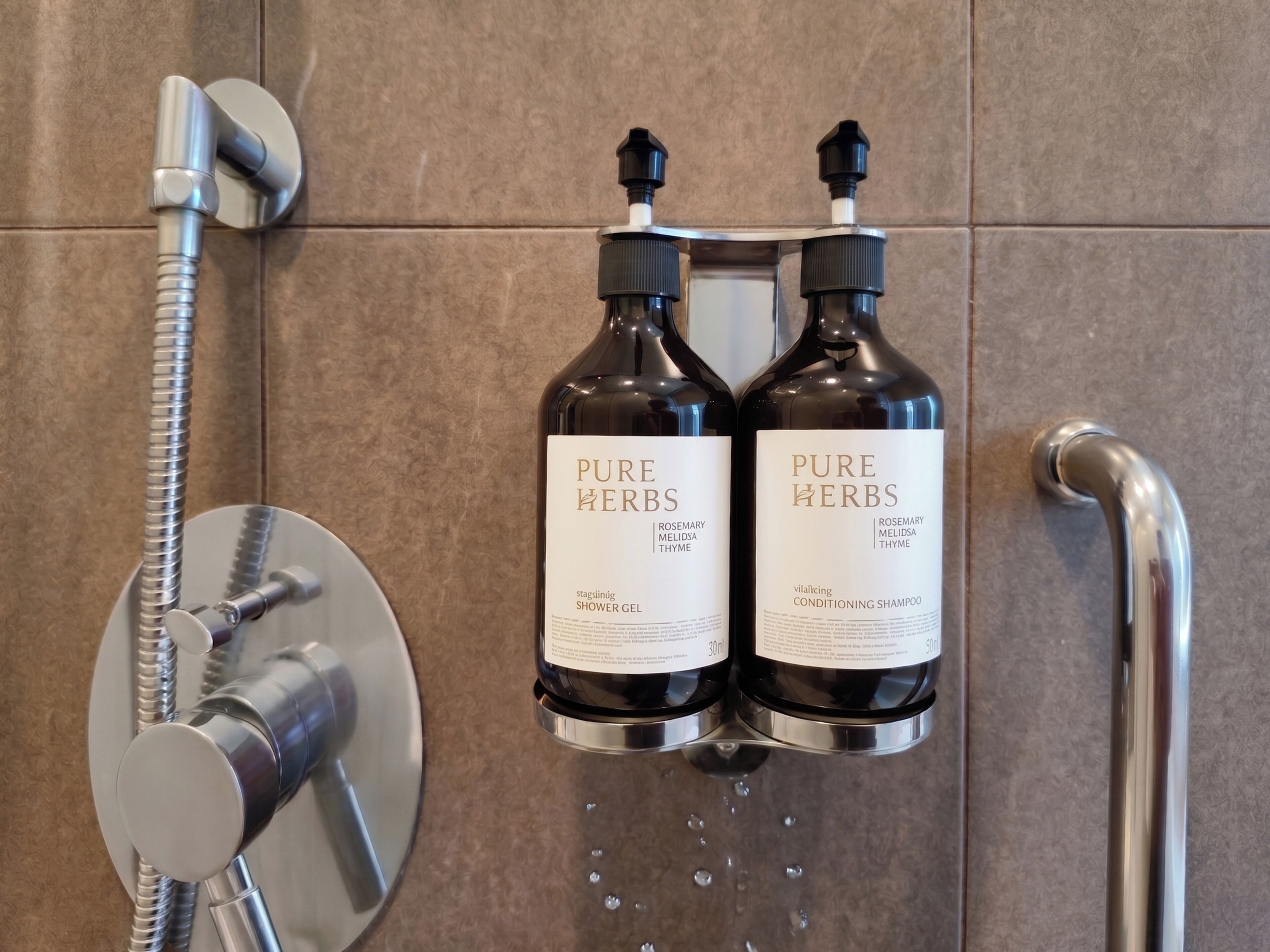 Zwei Flaschen Pure Herbs Duschgel und Conditioner-Shampoo sind an einer Wand neben einem Duschkopf angebracht.