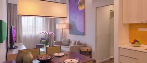 A hotel room with a dining area, a living area with a sofa and a TV, a large abstract painting, and a kitchen with fruit on the counter.
