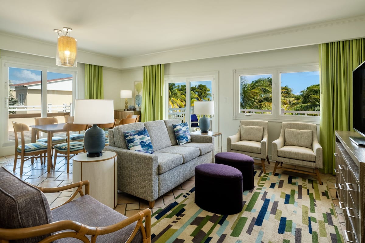 A spacious living room with a grey sofa, two armchairs, a coffee table, and a TV. The room has large windows with green curtains, a patterned rug, and a ceiling light fixture.