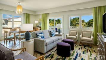 A spacious living room with a grey sofa, two armchairs, a coffee table, and a TV. The room has large windows with green curtains, a patterned rug, and a ceiling light fixture.