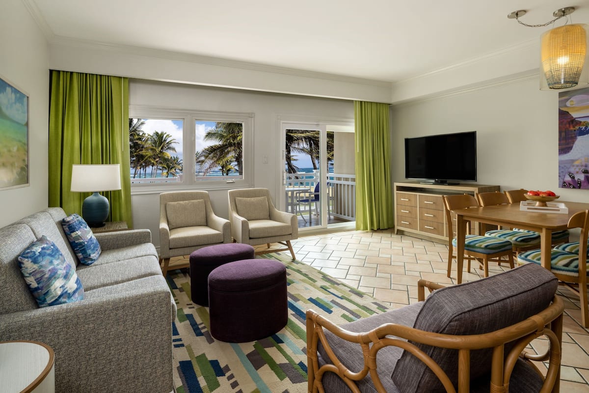 A hotel room with a sofa, armchairs, a dining table with chairs, a TV, and a balcony with a view of palm trees and the ocean.
