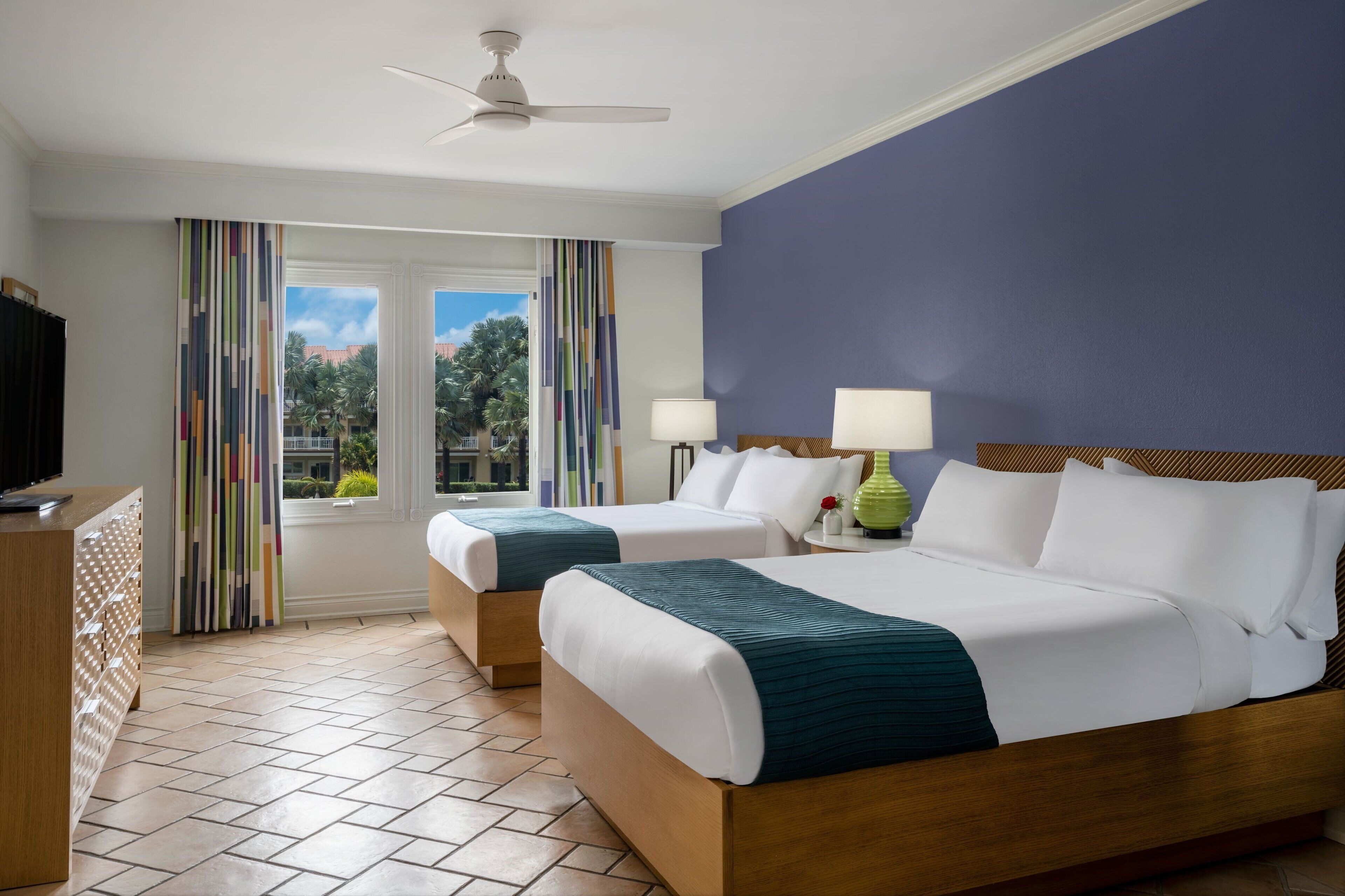 A hotel room with two beds, a TV, a ceiling fan, and a window with curtains.