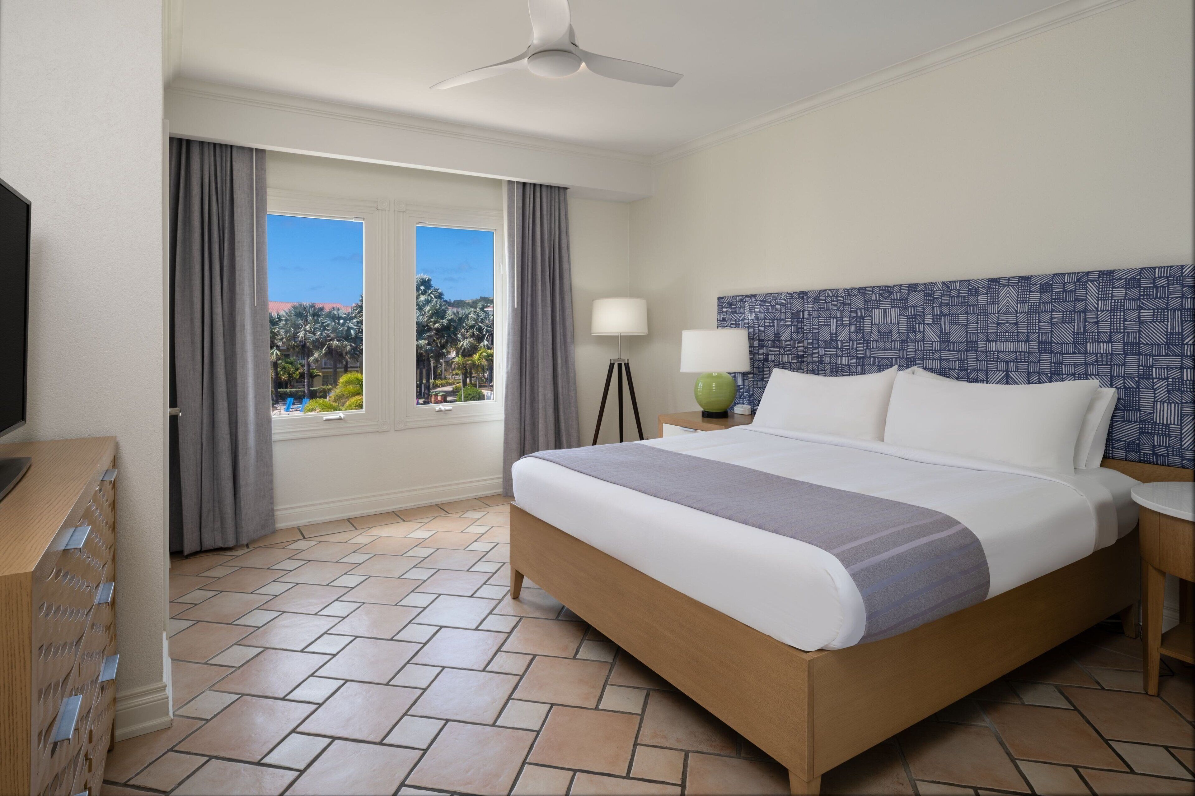 A hotel room with a bed, a television, a desk with drawers, a lamp, and a view of palm trees.