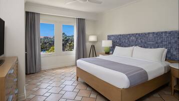 A hotel room with a bed, a television, a desk with drawers, a lamp, and a view of palm trees.