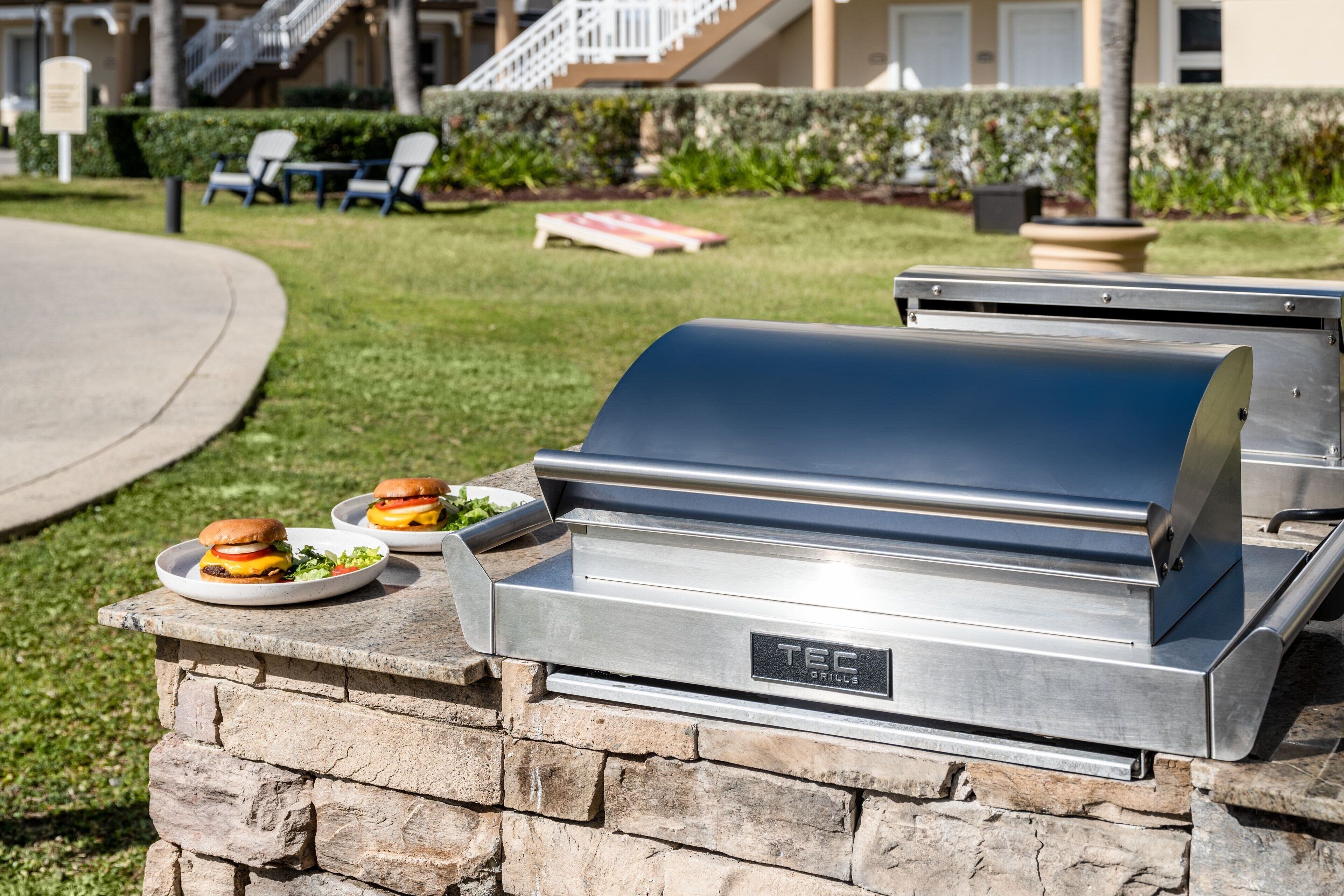 A stone barbecue grill with two burgers on plates, set against a backdrop of a residential area with a staircase and outdoor seating.