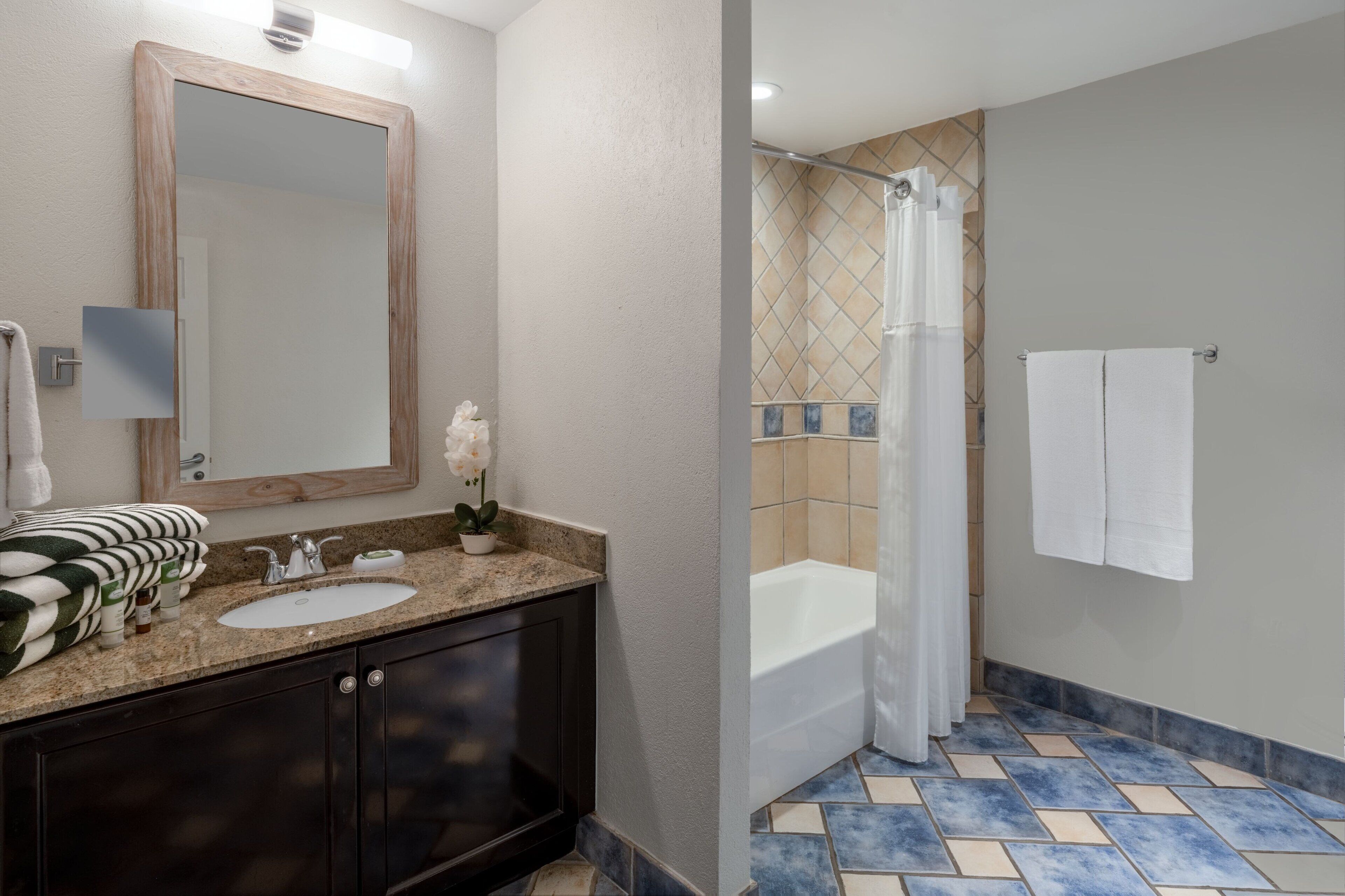 A bathroom with a wooden-framed mirror, a sink with a granite countertop, and a shower with a tiled floor.