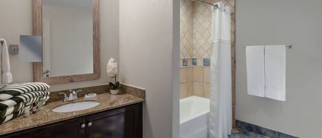 Un baño con espejo de marco de madera, lavabo con encimera de granito y ducha con suelo de baldosas.