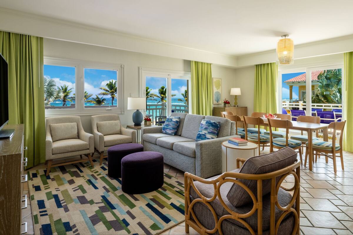 A hotel room with a sofa, chairs, a dining table, and a view of the ocean.