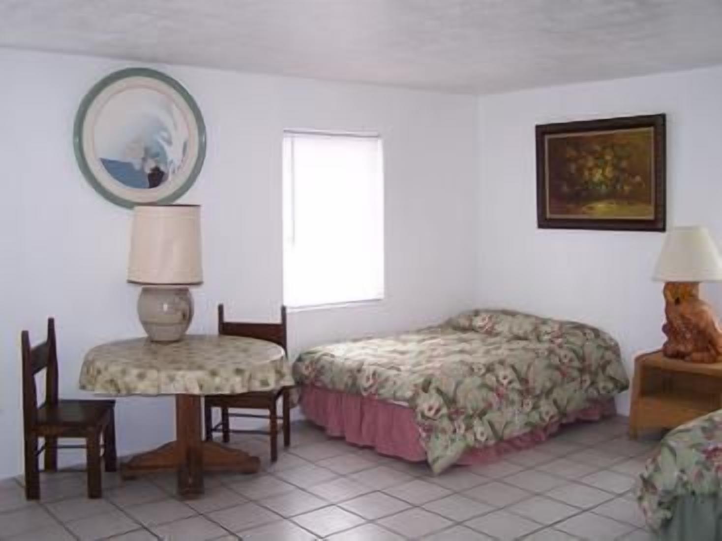 Een kamer met een bed, een tafel met stoel, een lamp, een raam en een schilderij aan de muur.