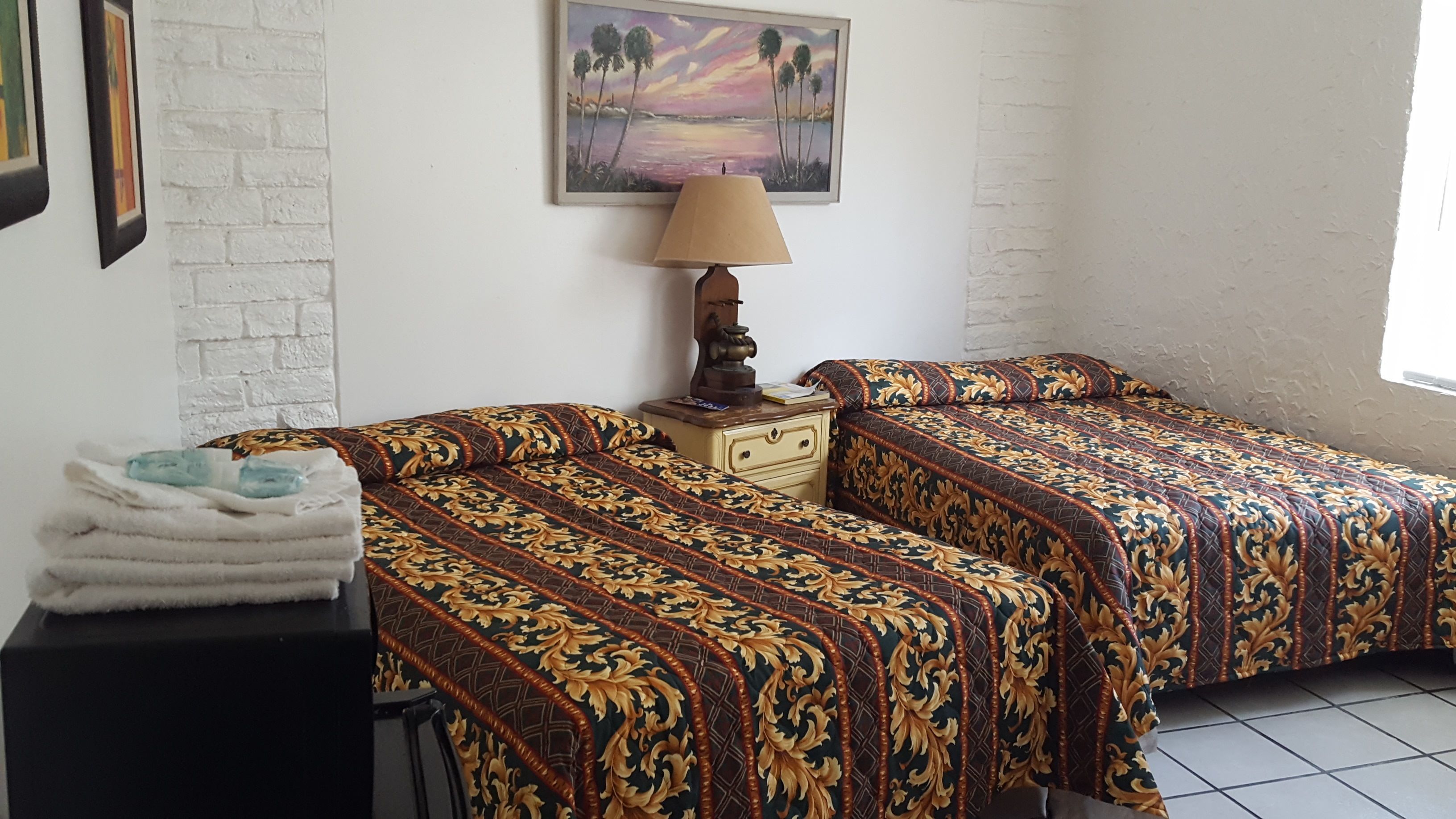 A room with two beds, a nightstand with a lamp, a painting on the wall, and a stack of towels on a table.