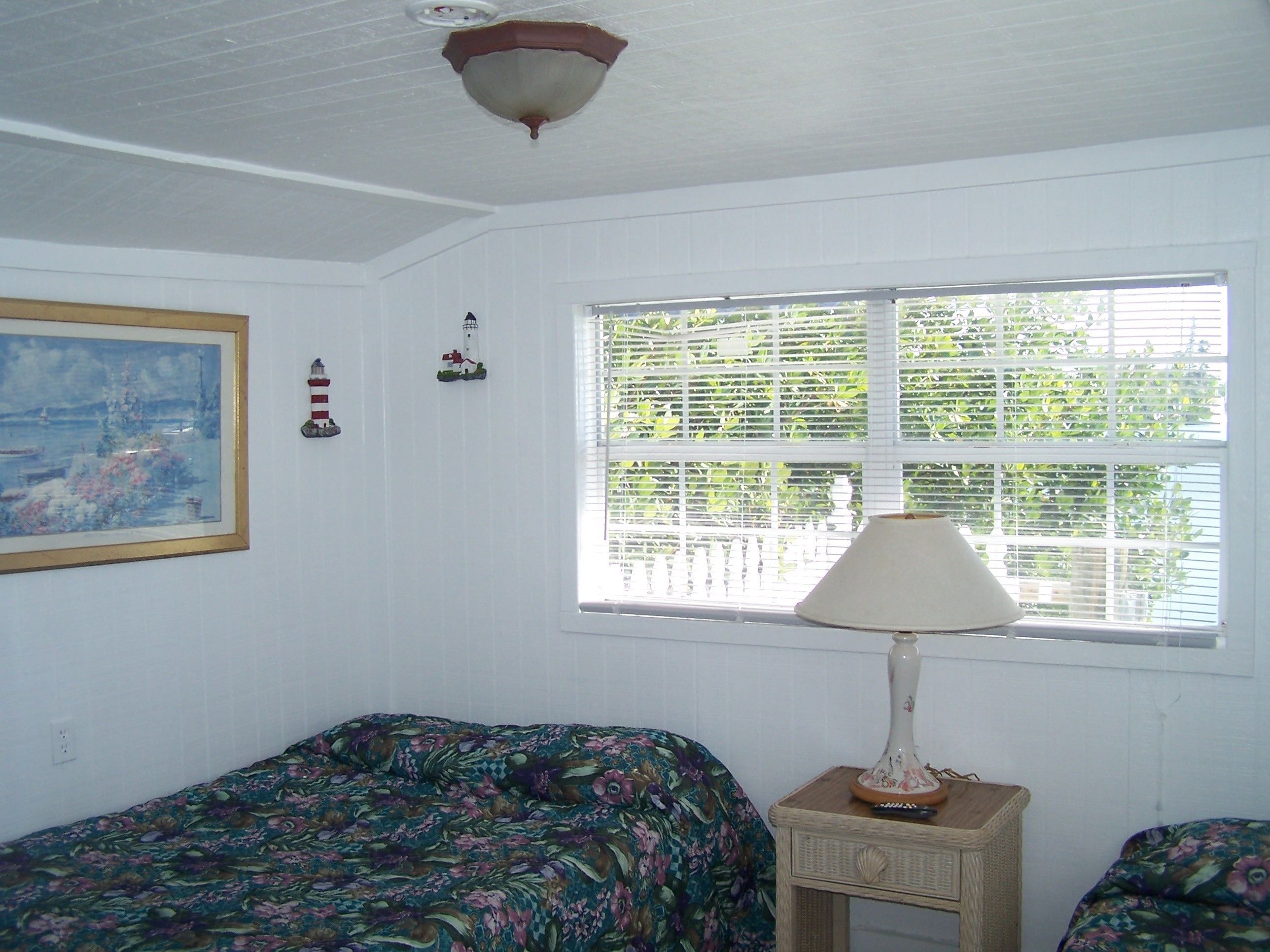 A room with a bed, a nightstand, a lamp, a window with blinds, and a painting on the wall.
