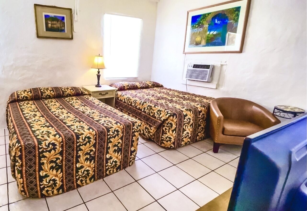 A room with two beds, a chair, a desk, a lamp, and a TV.