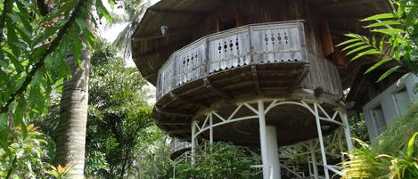 A wooden structure with a balcony, surrounded by lush greenery and a stone pathway.