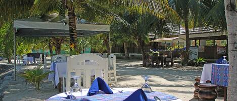 An outdoor dining area with tables set for two, surrounded by palm trees and a sandy ground.
