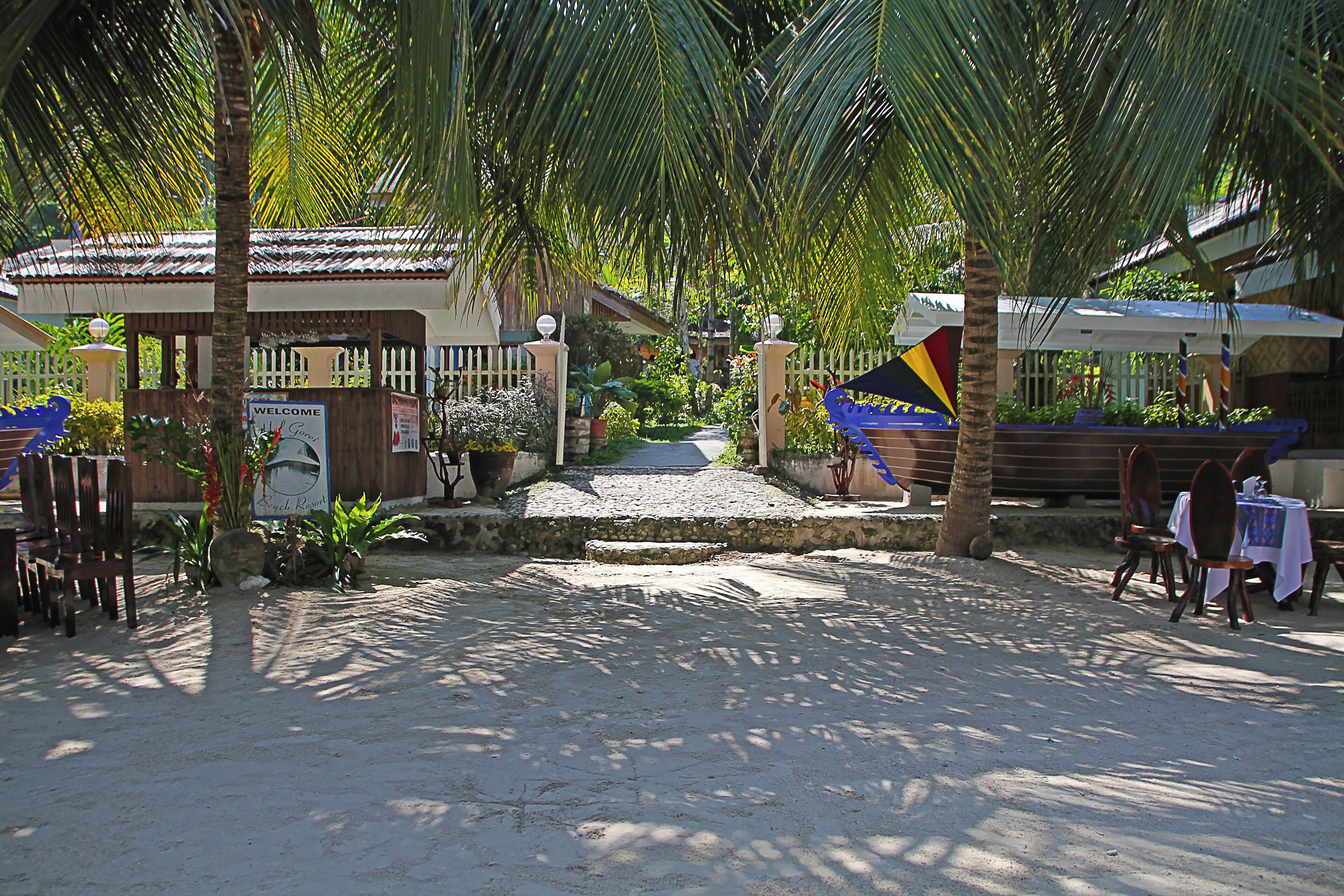 Entrada a un resort tropical con un letrero de bienvenida, palmeras y un área de descanso cubierta.