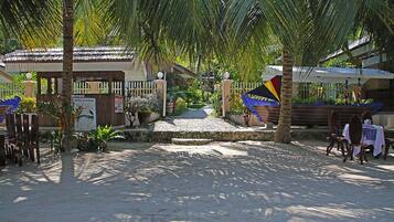 Entrada a un resort tropical con un letrero de bienvenida, palmeras y un área de descanso cubierta.