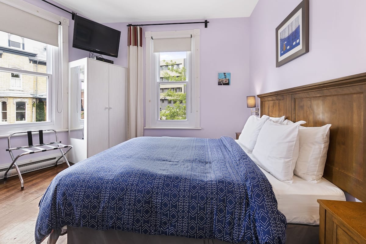 A bedroom with a bed, a TV mounted on the wall, a window with a view of buildings, and a door.