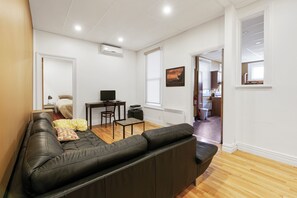A modern living room with a leather sofa, a small table, and a television. There is a window with a view of the outside, and a door leading to another room. - Château des Tourelles - Par Aneyro (Québec City)