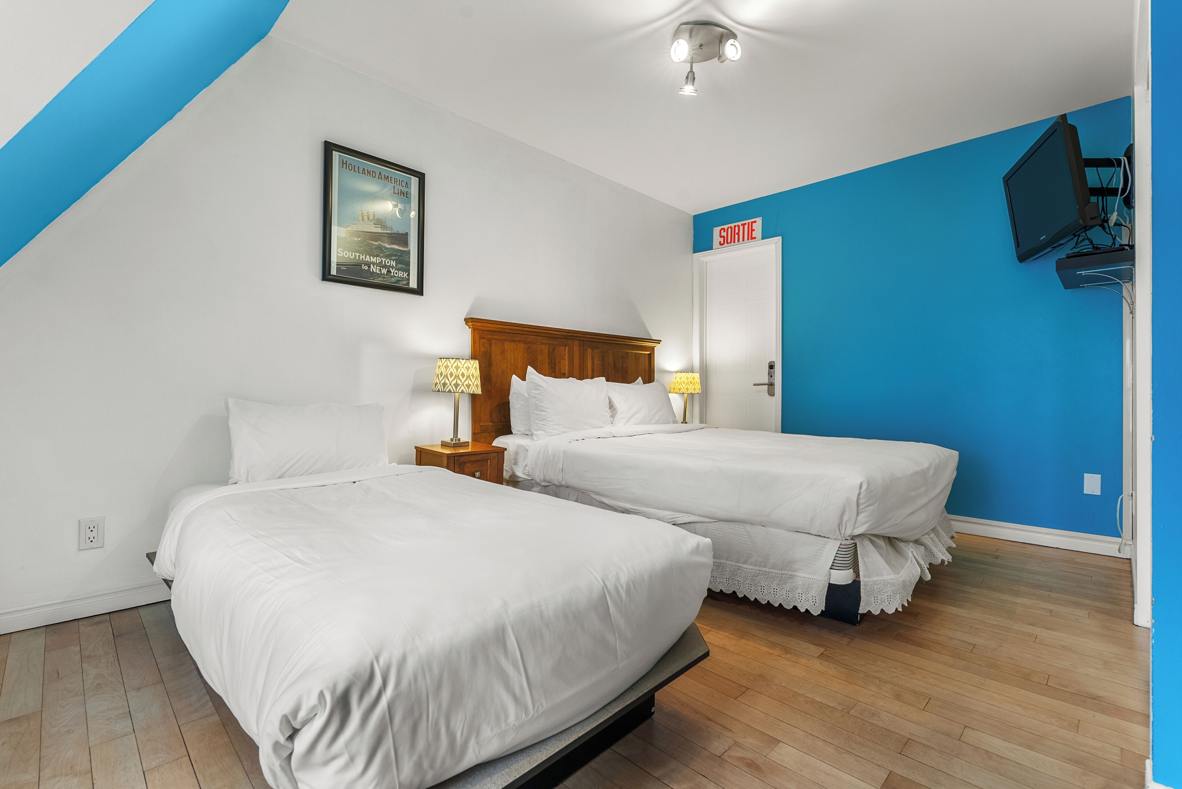 A hotel room with two beds, a wooden headboard, a framed picture on the wall, a blue accent wall, and a television mounted on the wall.