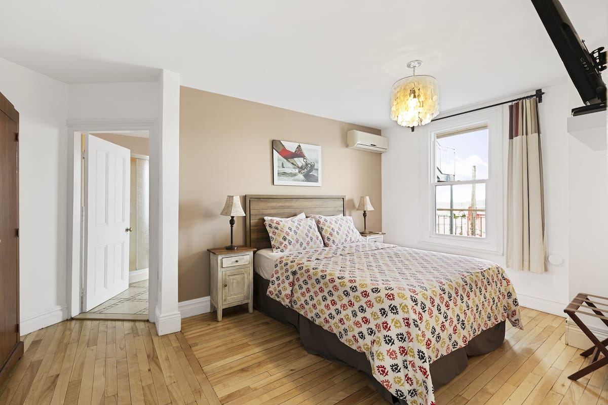 A bedroom with a bed, nightstands, a chandelier, a window with curtains, and a door leading to another room.