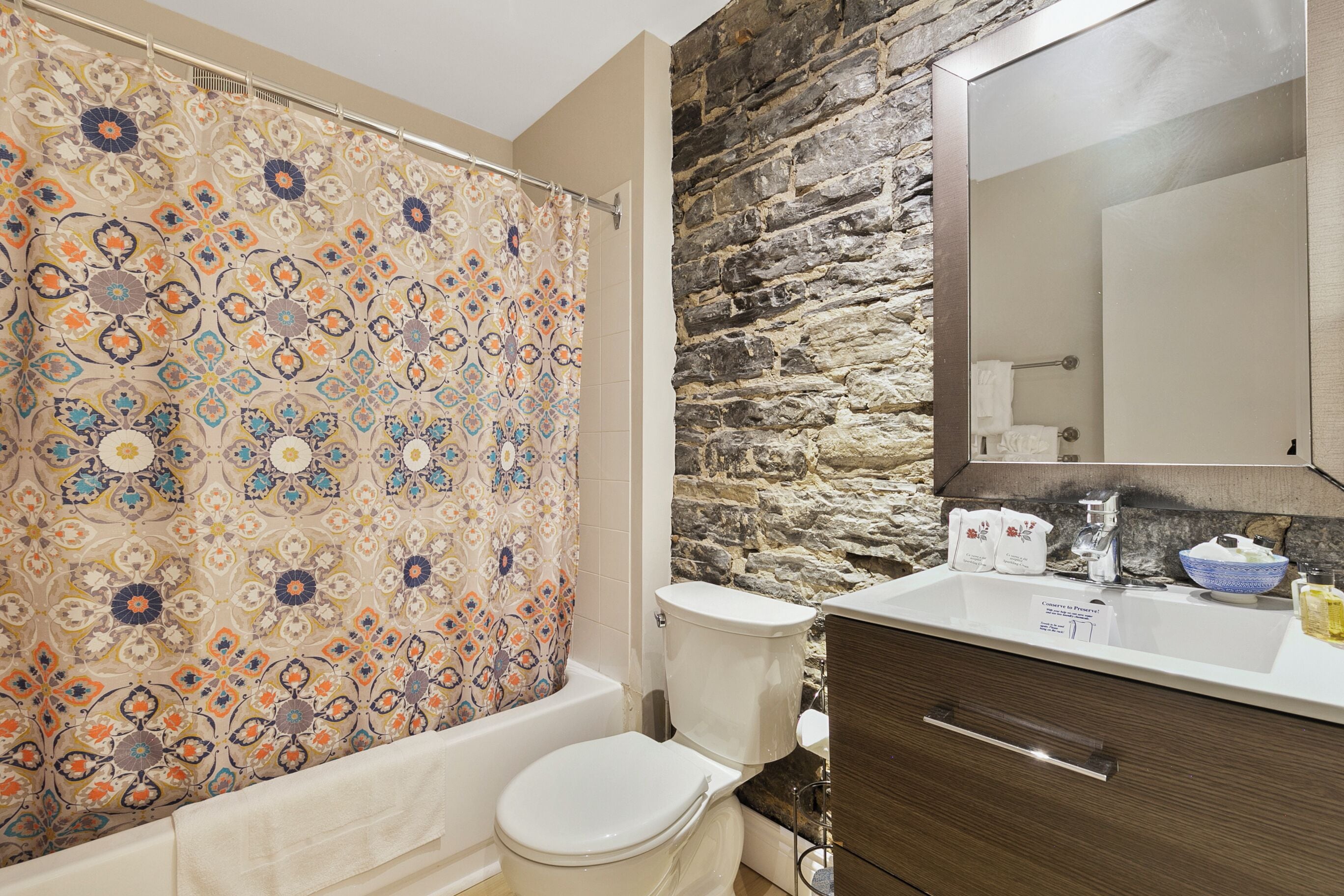 A bathroom with a patterned shower curtain, a toilet, a sink with a mirror, and a stone wall.