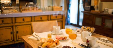 A breakfast table with croissants, fruit, and orange juice in a cozy room with wooden furniture and a kitchen in the background.