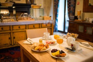 Une table de petit-déjeuner avec des croissants, des fruits et du jus d’orange, dans une pièce chaleureuse aux meubles en bois, avec une cuisine en arrière-plan.